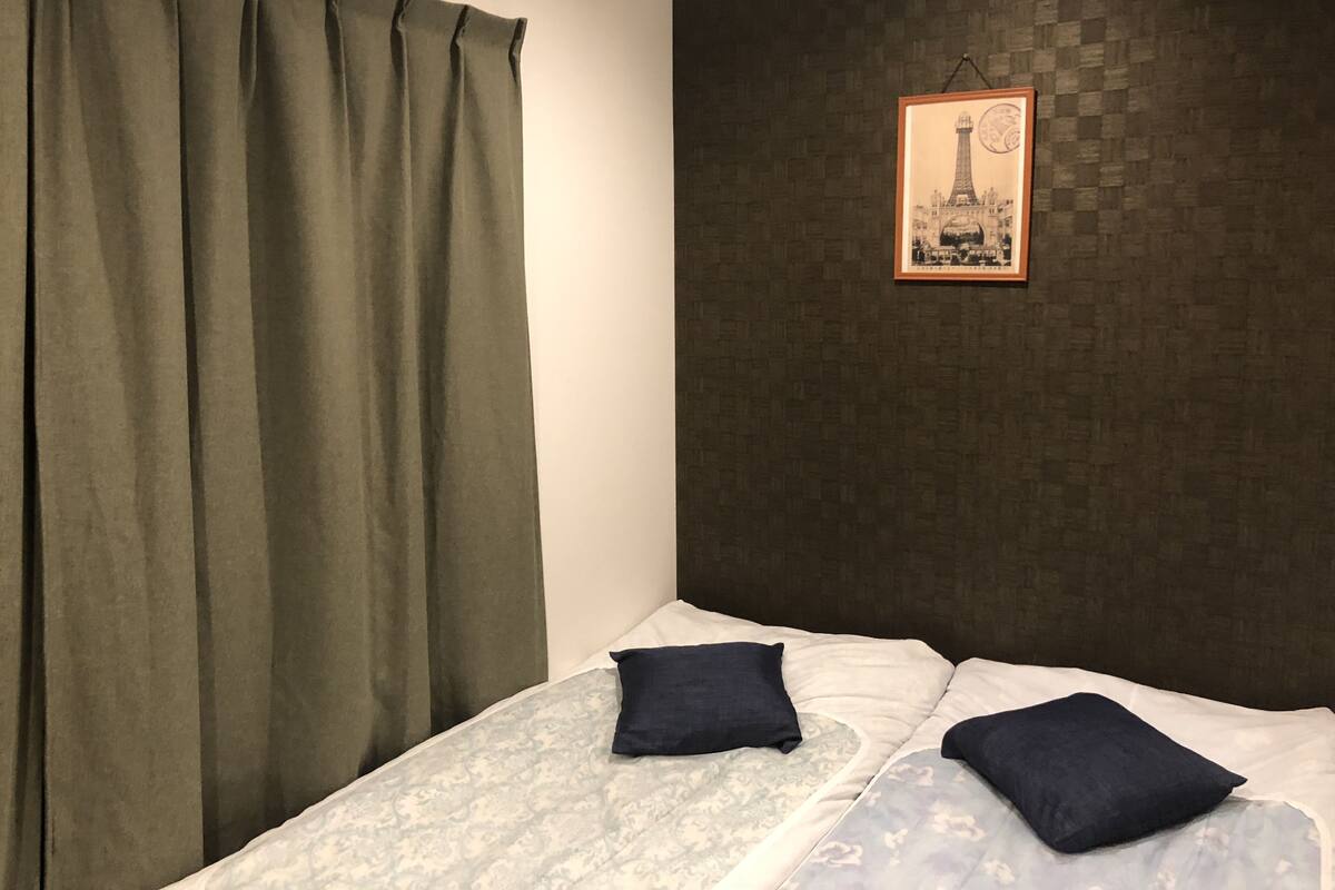 A bed with a patterned bedspread and two pillows, a dark wall with a framed picture, and a green curtain.