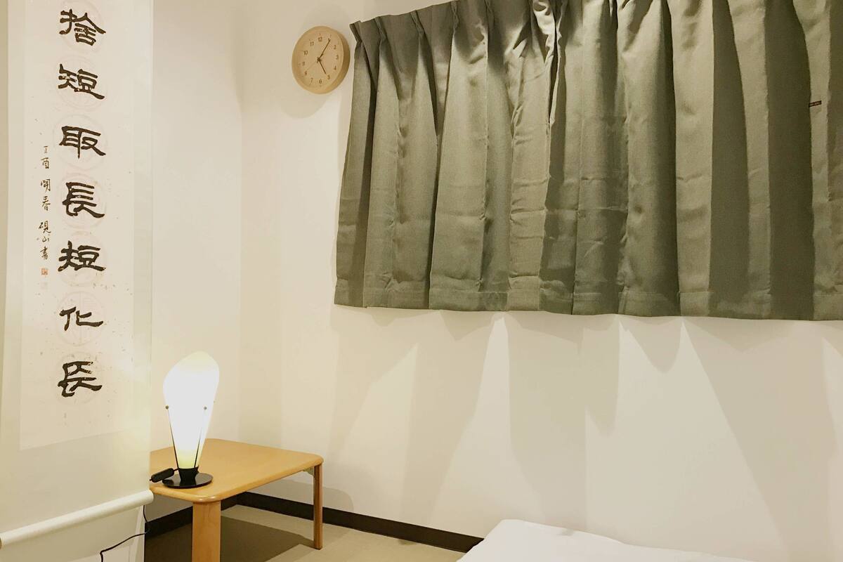 A room with a bed, a bedside table with a lamp, a wall clock, and a curtain.