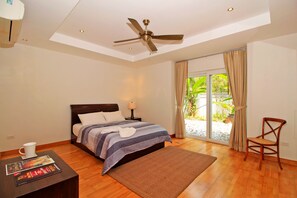 A bedroom with a bed, a ceiling fan, a nightstand, a coffee table, and a view of the outdoors.