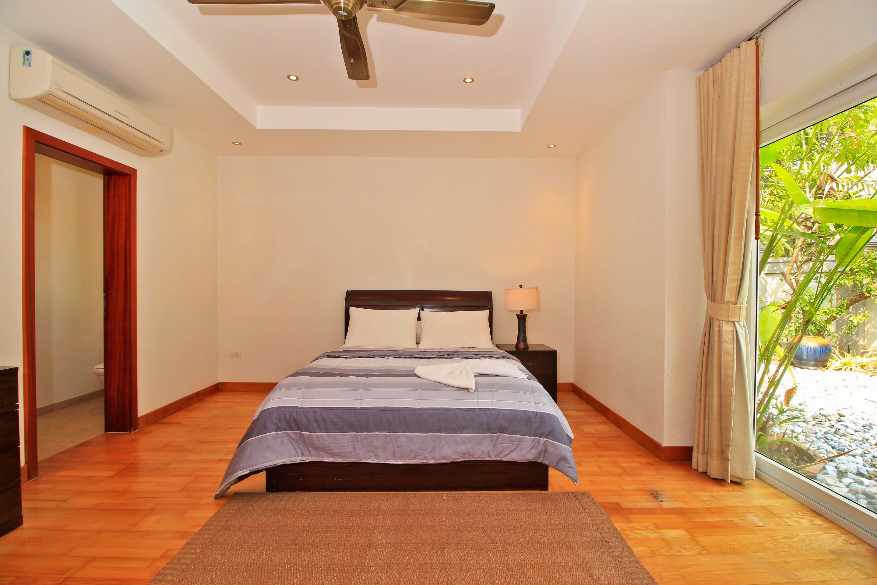A bedroom with a bed, a ceiling fan, a nightstand with a lamp, a window with curtains, and a view of greenery outside.
