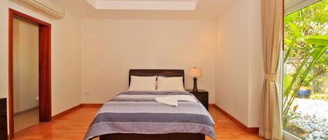 A bedroom with a bed, a ceiling fan, a nightstand with a lamp, a window with curtains, and a view of greenery outside.