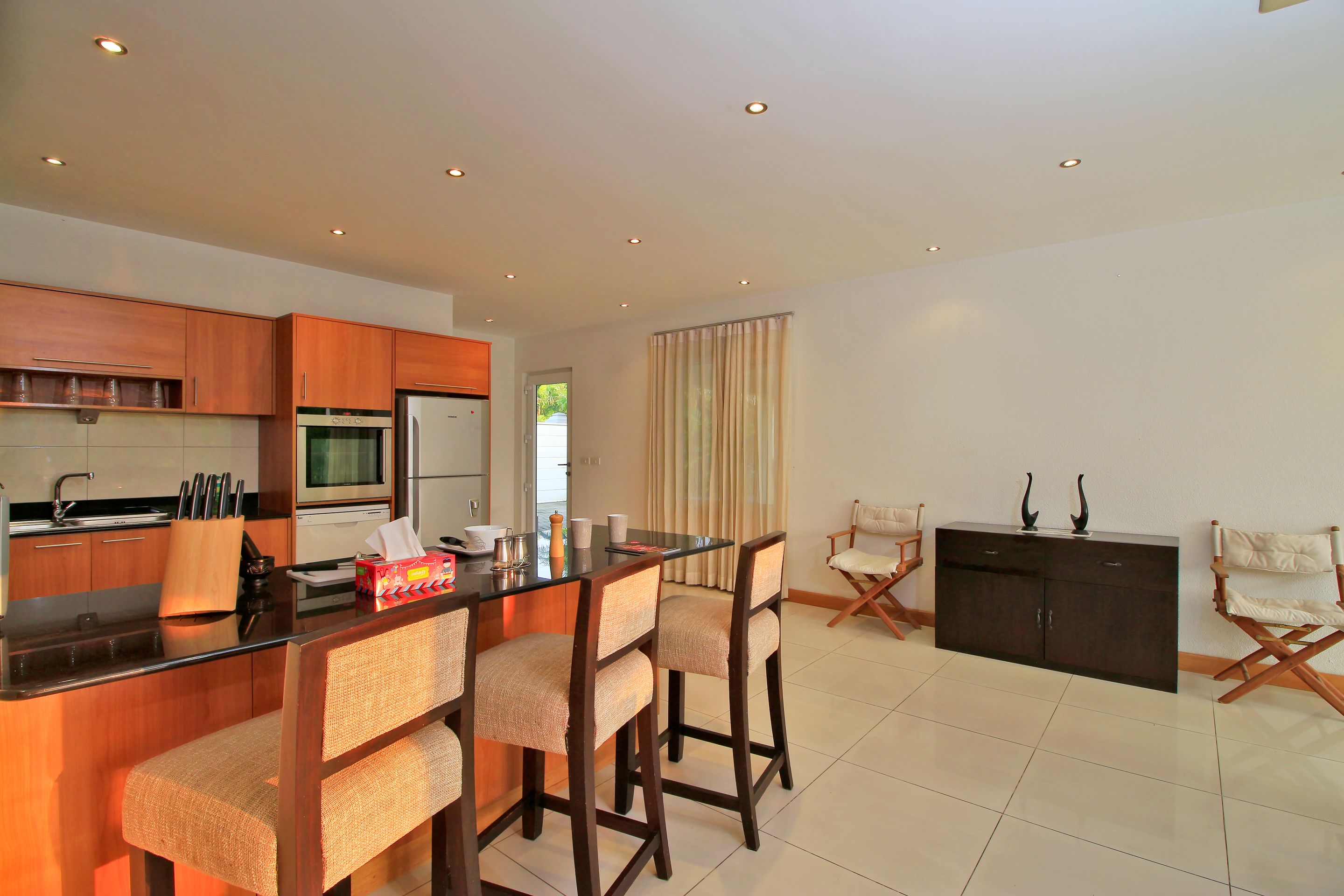 A modern kitchen with wooden cabinets, a central island with bar stools, a refrigerator, and a dining area with a dark cabinet and two chairs.