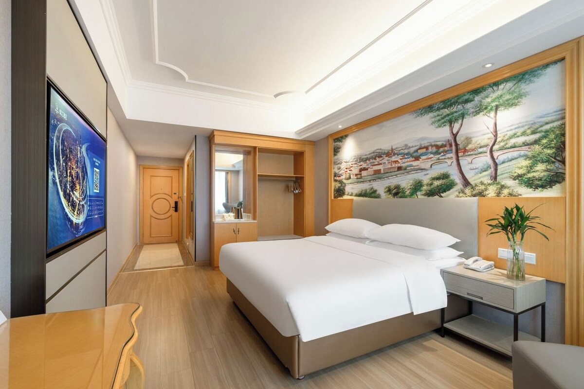 A hotel room with a bed, a desk, a TV, a painting, and a mirror.