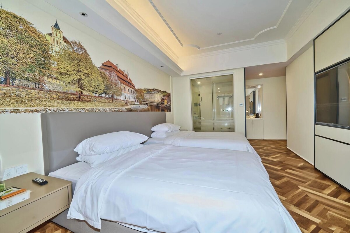 A hotel room with a large bed, a nightstand, a painting of a town scene, a mirror, and a TV.