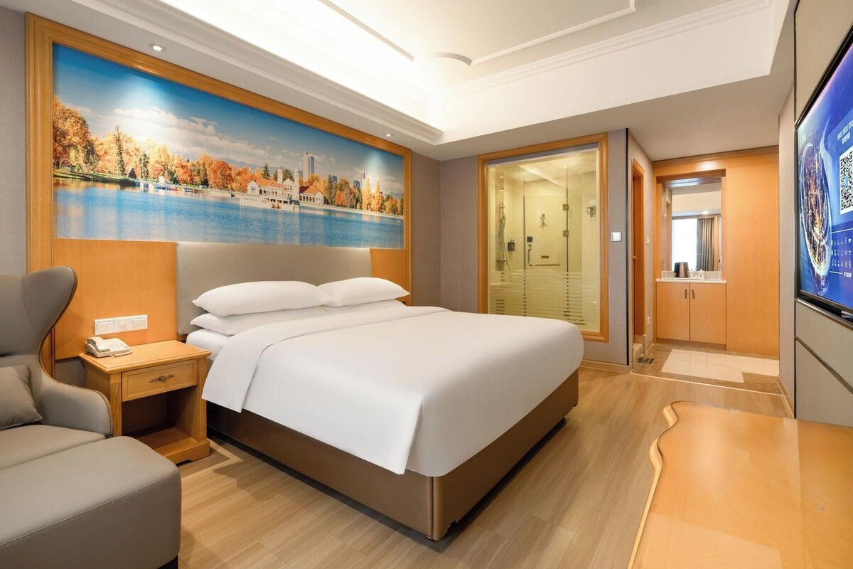A hotel room with a bed, a nightstand, a chair, a painting of a cityscape, and a bathroom with a glass door.
