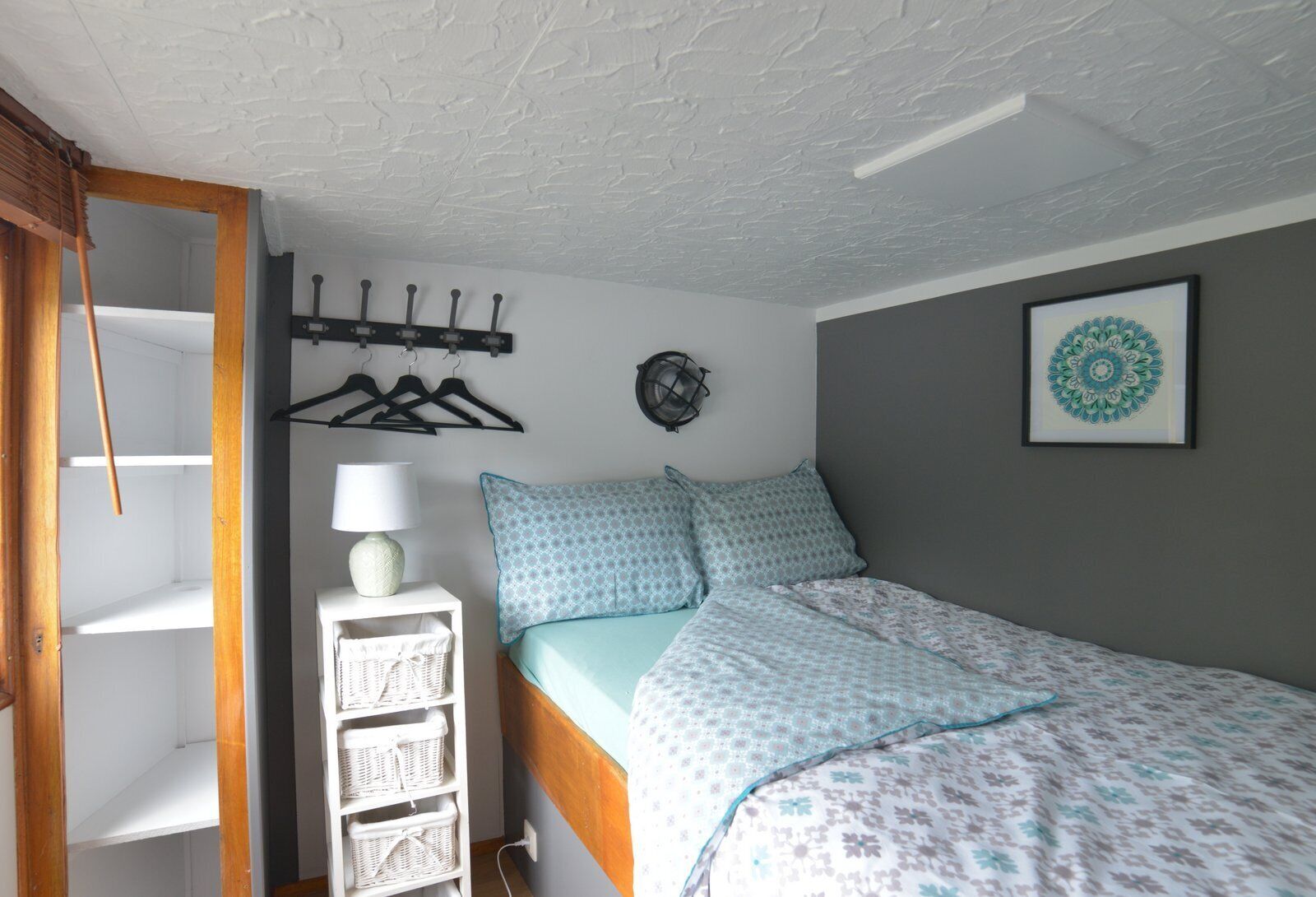 A bedroom with a bed, a wall-mounted lamp, a framed artwork, and a white shelf with baskets.