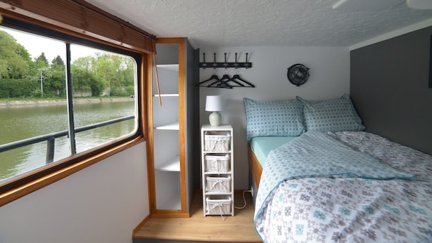 Un dormitorio con cama, ventana con vista al río, estantería blanca con cestas y perchero de pared.