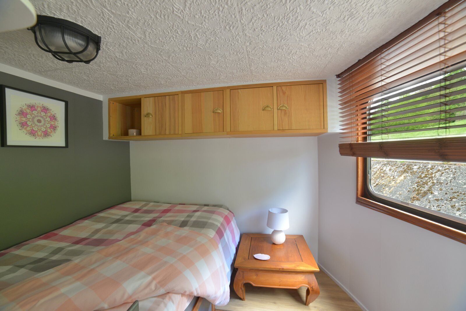 Un dormitorio con una cama, un armario de madera sobre la cama, una mesita de noche de madera con una lámpara y una ventana con persianas.