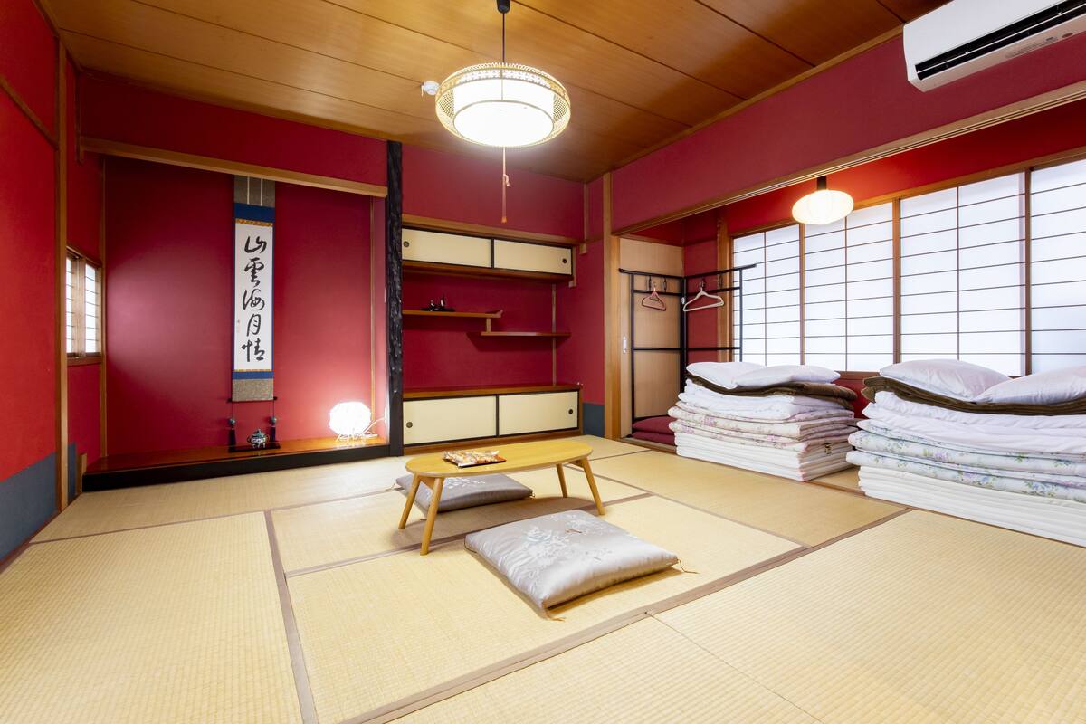 A traditional Japanese room with tatami flooring, a low wooden table, a hanging light fixture, and a stack of neatly folded bedding.