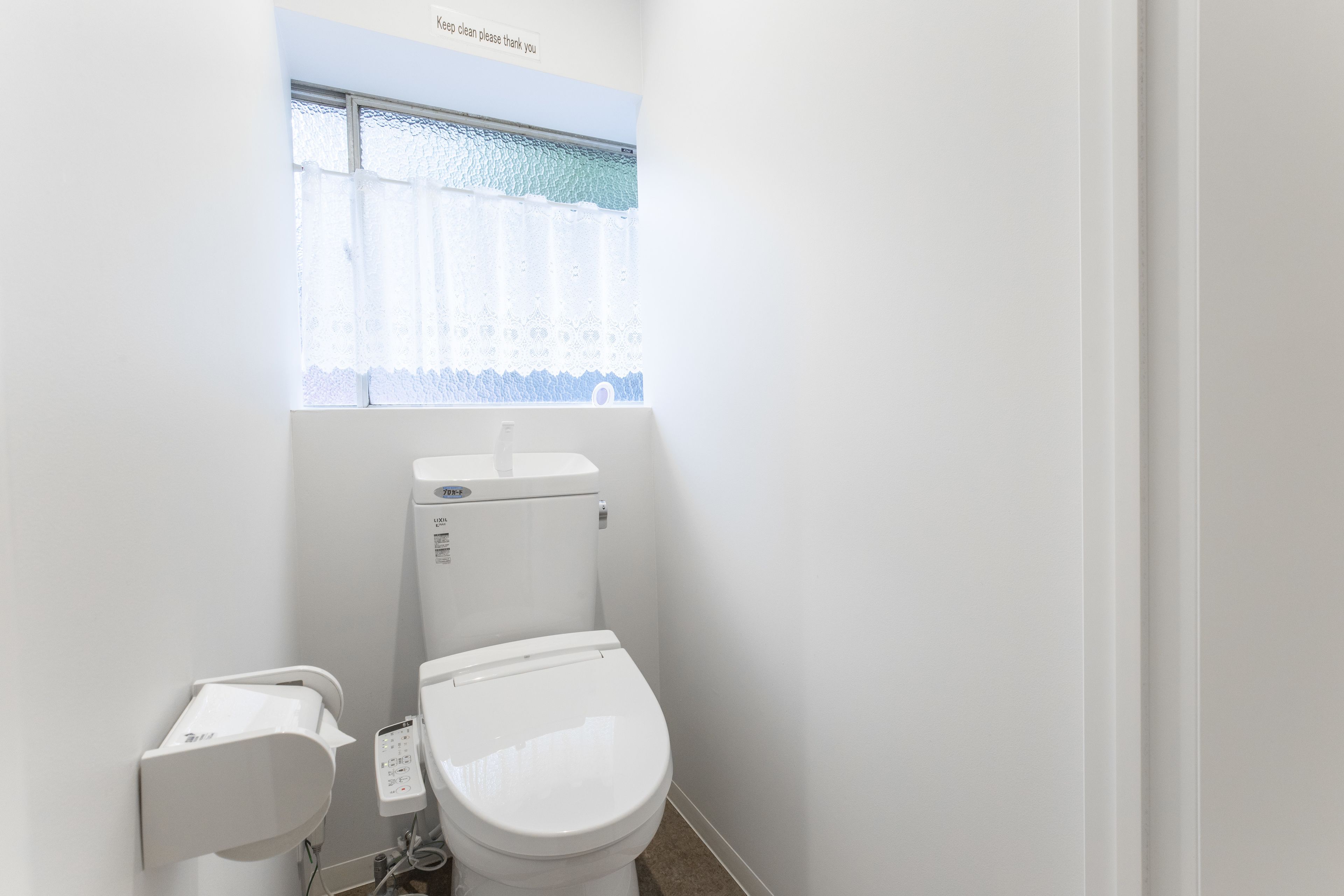 A small, clean bathroom with a toilet, a wall-mounted bidet, and a window with a lace curtain.