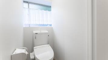 A small, clean bathroom with a toilet, a wall-mounted bidet, and a window with a lace curtain.
