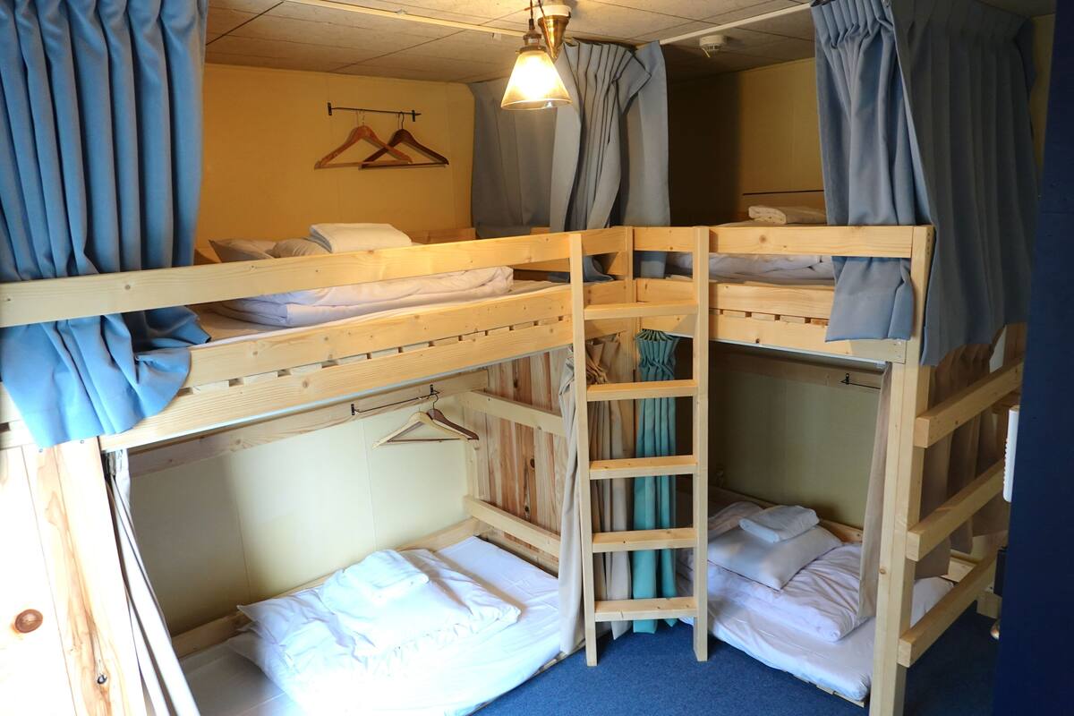 A bunk bed room with a wooden ladder, a hanging clothes rack, and a single bed with a white mattress.