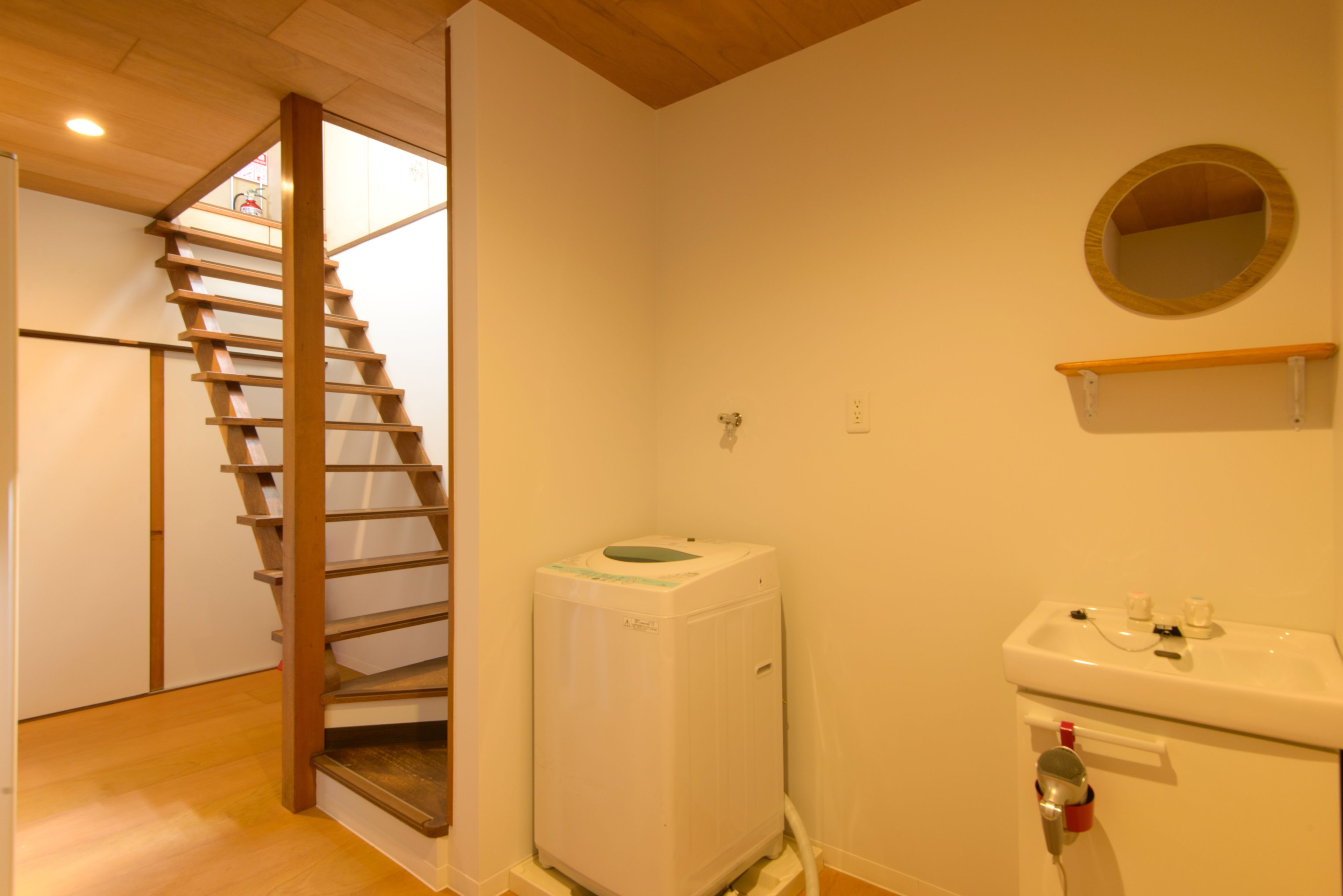 A bathroom with a wooden staircase, a white washing machine, a sink with a mirror, and a shelf above the sink.