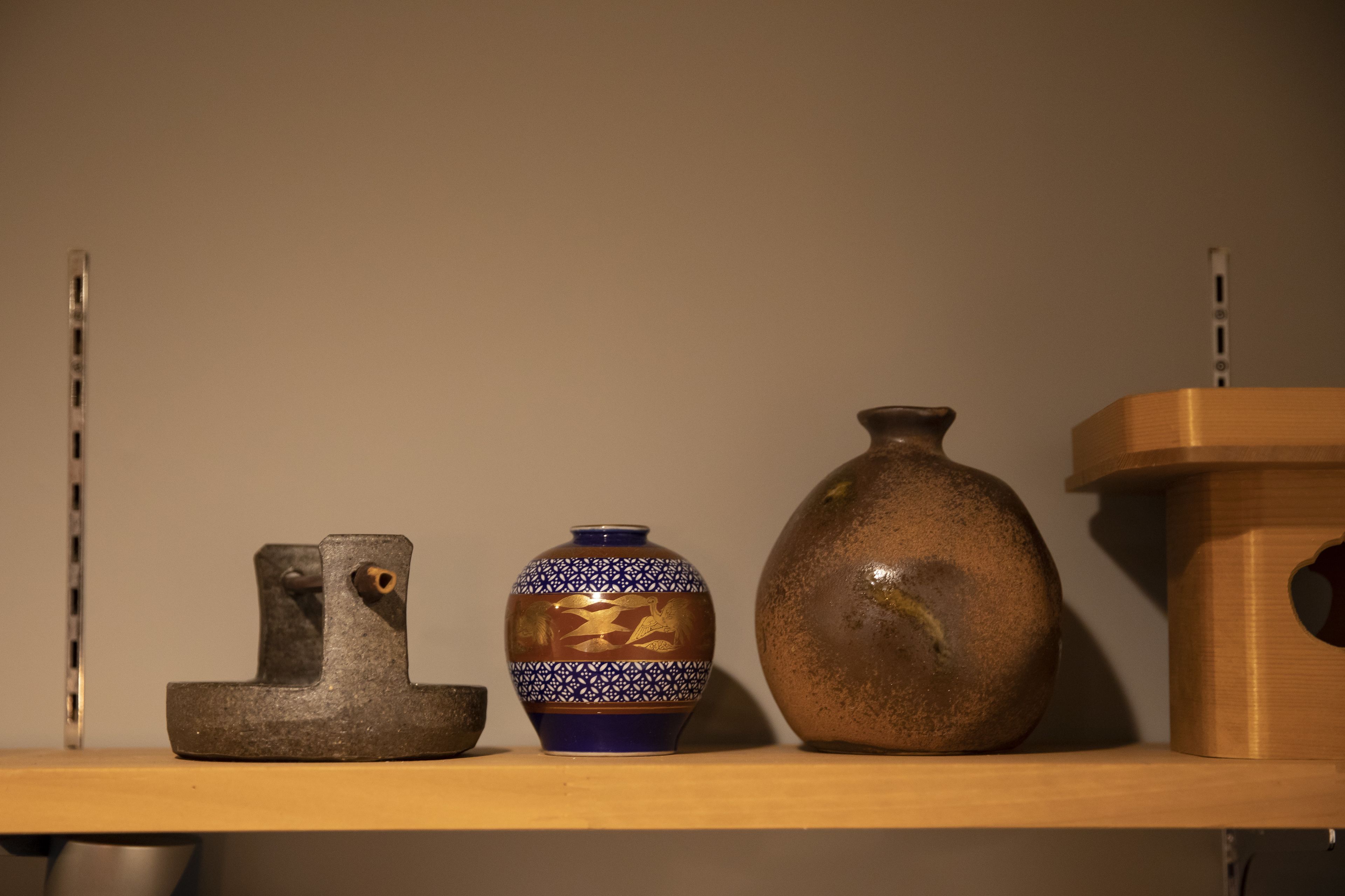 Uma prateleira com três objetos de cerâmica: uma escultura pequena, escura e geométrica; um vaso de tamanho médio com padrões azuis e dourados; e um vaso grande, marrom e texturizado.