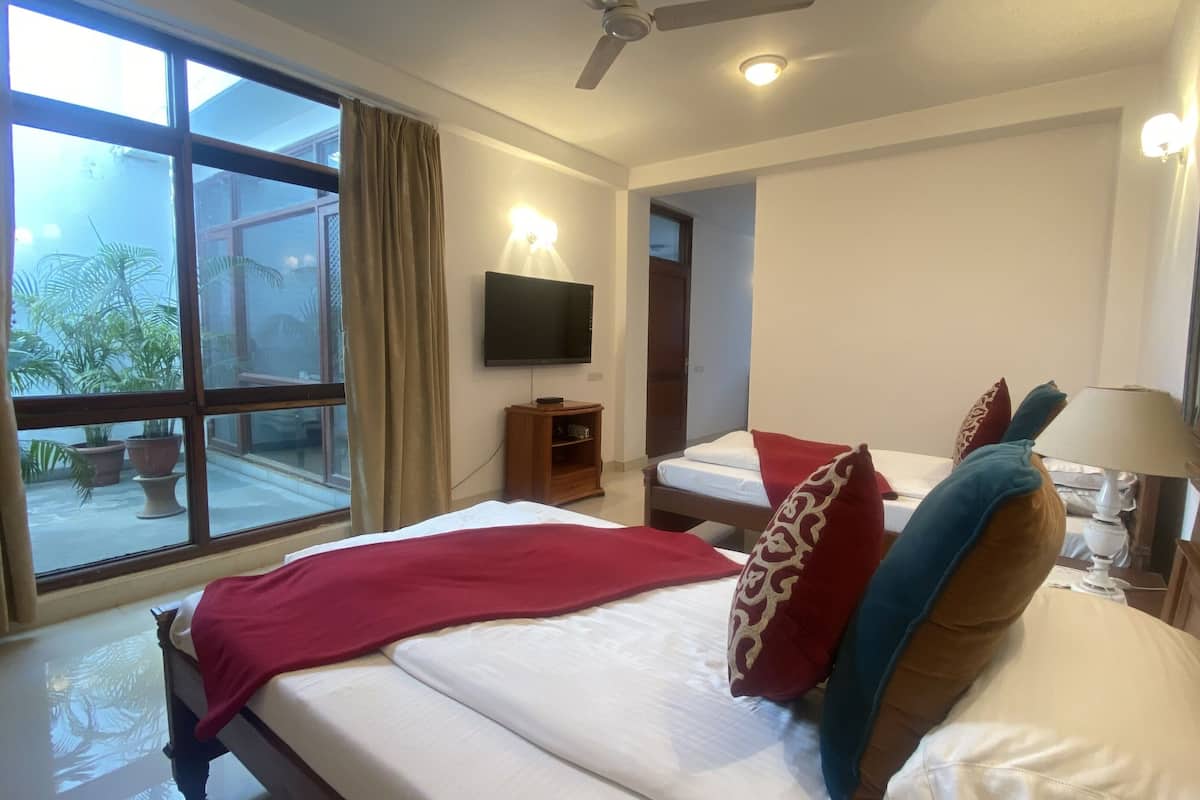 A hotel room with a large window, a bed with red and blue pillows, a bedside table with a lamp, and a TV on the wall.