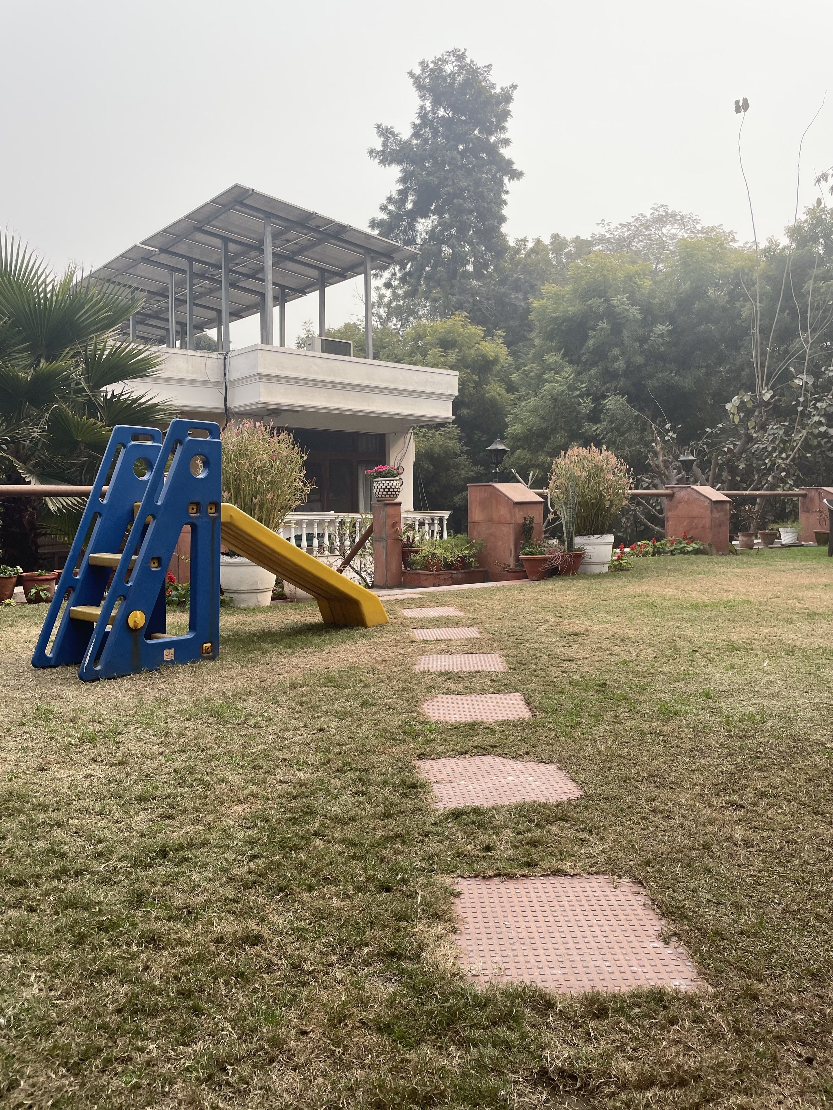 A playground with a slide and climbing structure, surrounded by potted plants and a building with a covered area.