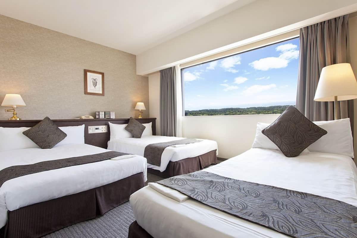 A hotel room with two beds, a large window with a view, a bedside table with a lamp, and a framed picture on the wall.