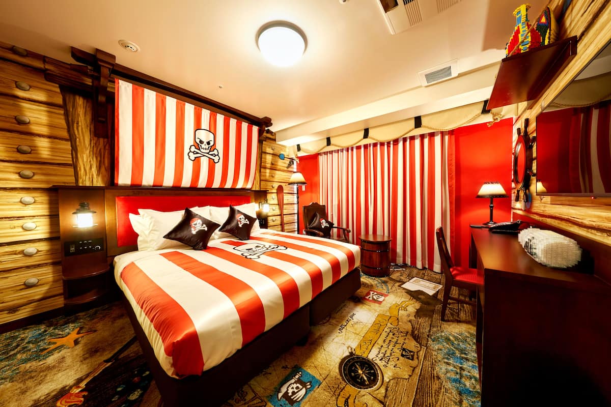 A room with a bed featuring a striped headboard, a desk, a chair, and a red curtain.