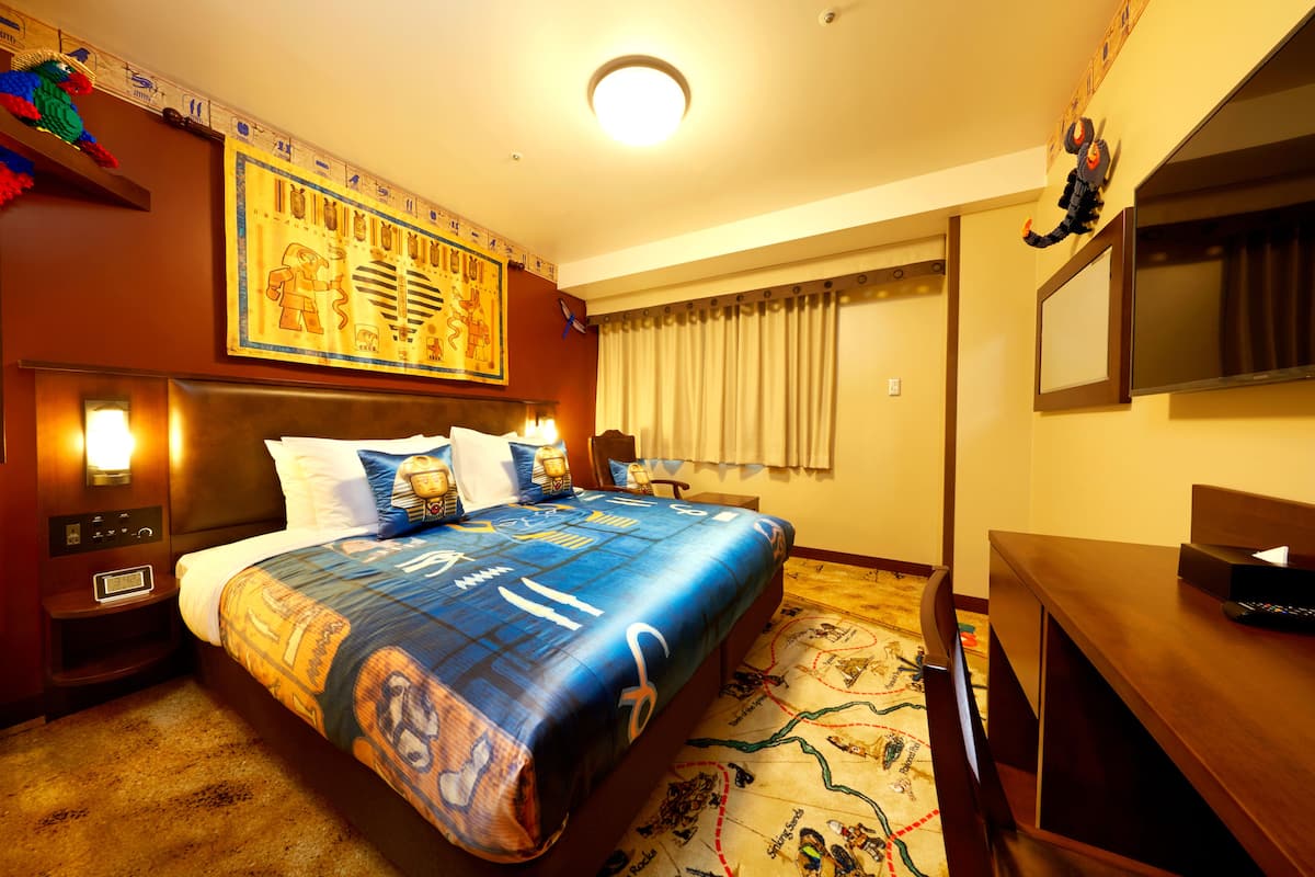 A hotel room with a large bed, a desk, a chair, a nightstand, a wall-mounted TV, and a window with curtains.