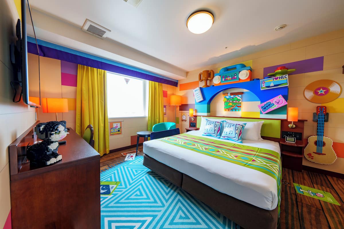A hotel room with a bed, a desk, a chair, a guitar, and a TV.