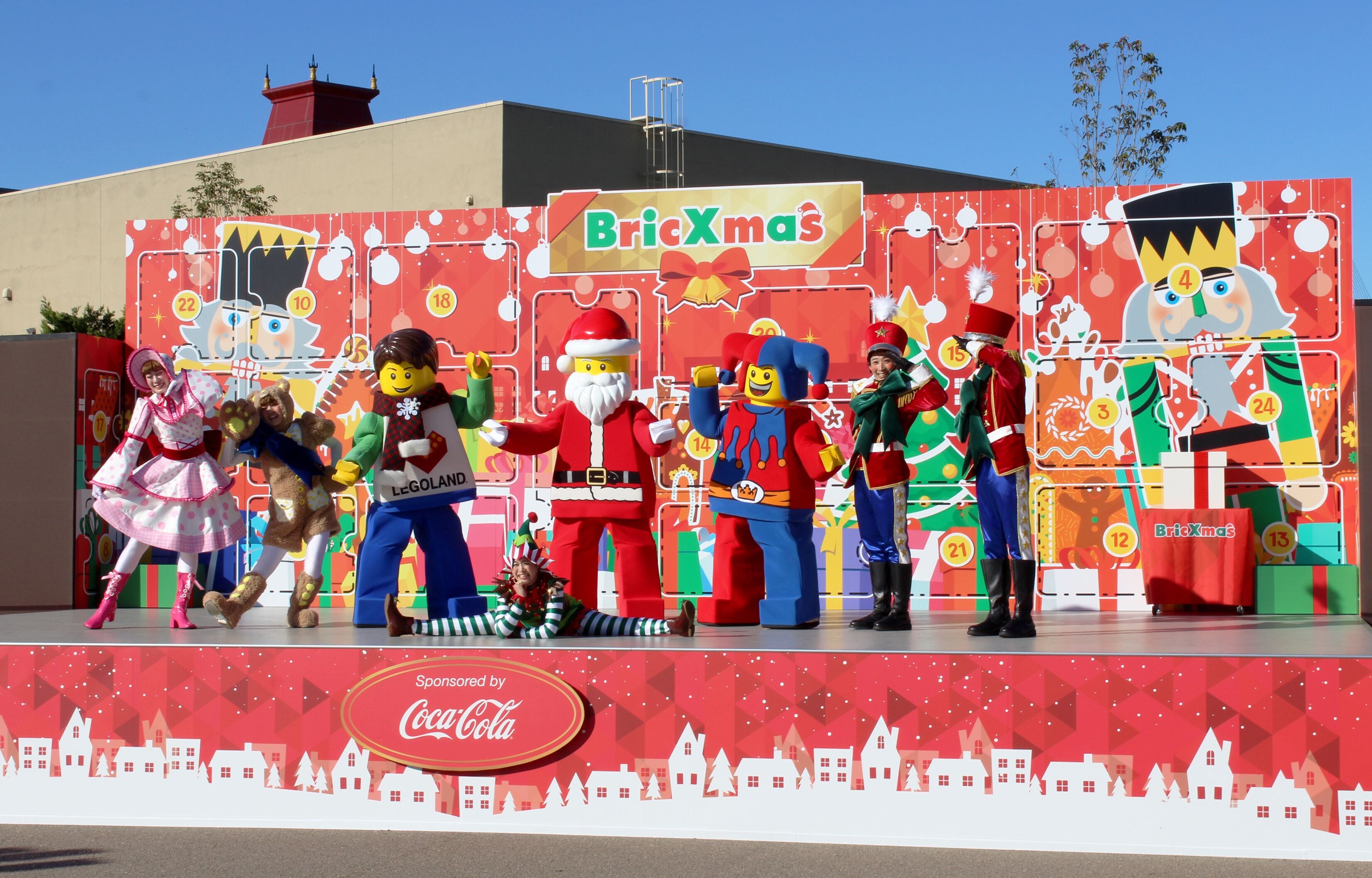 A stage with LEGO characters in festive costumes, sponsored by Coca-Cola, with a backdrop featuring a Christmas theme and the text "BricXmas".