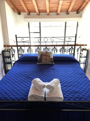 A bed with a blue quilted bedspread, a decorative pillow, and two folded white towels.