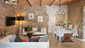 Un salon chaleureux avec un mur en pierre, une cheminée, un canapé blanc et un coin repas avec une table mise pour quatre personnes.