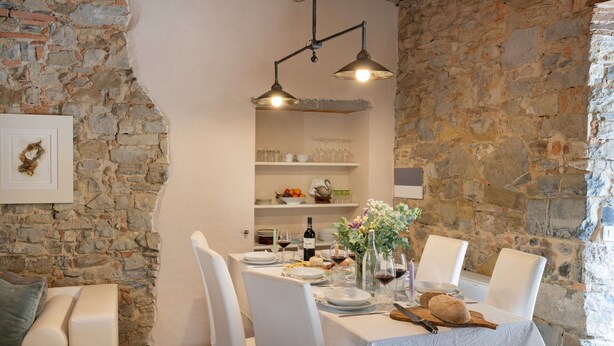 Un coin repas avec un mur en pierre, une table blanche dressée pour quatre personnes, et une étagère avec de la verrerie et de la vaisselle en arrière-plan.