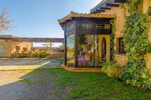 A glass-enclosed structure with a patio, surrounded by greenery and a stone building.