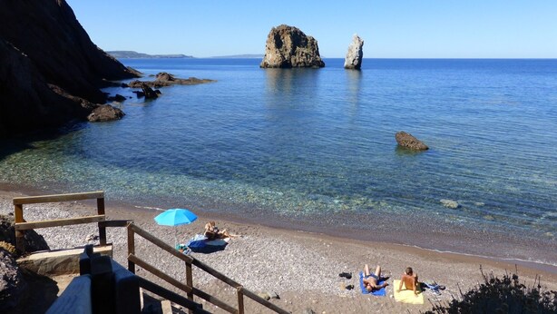 A beach with clear water, rocky formations, and a wooden deck with umbrellas.