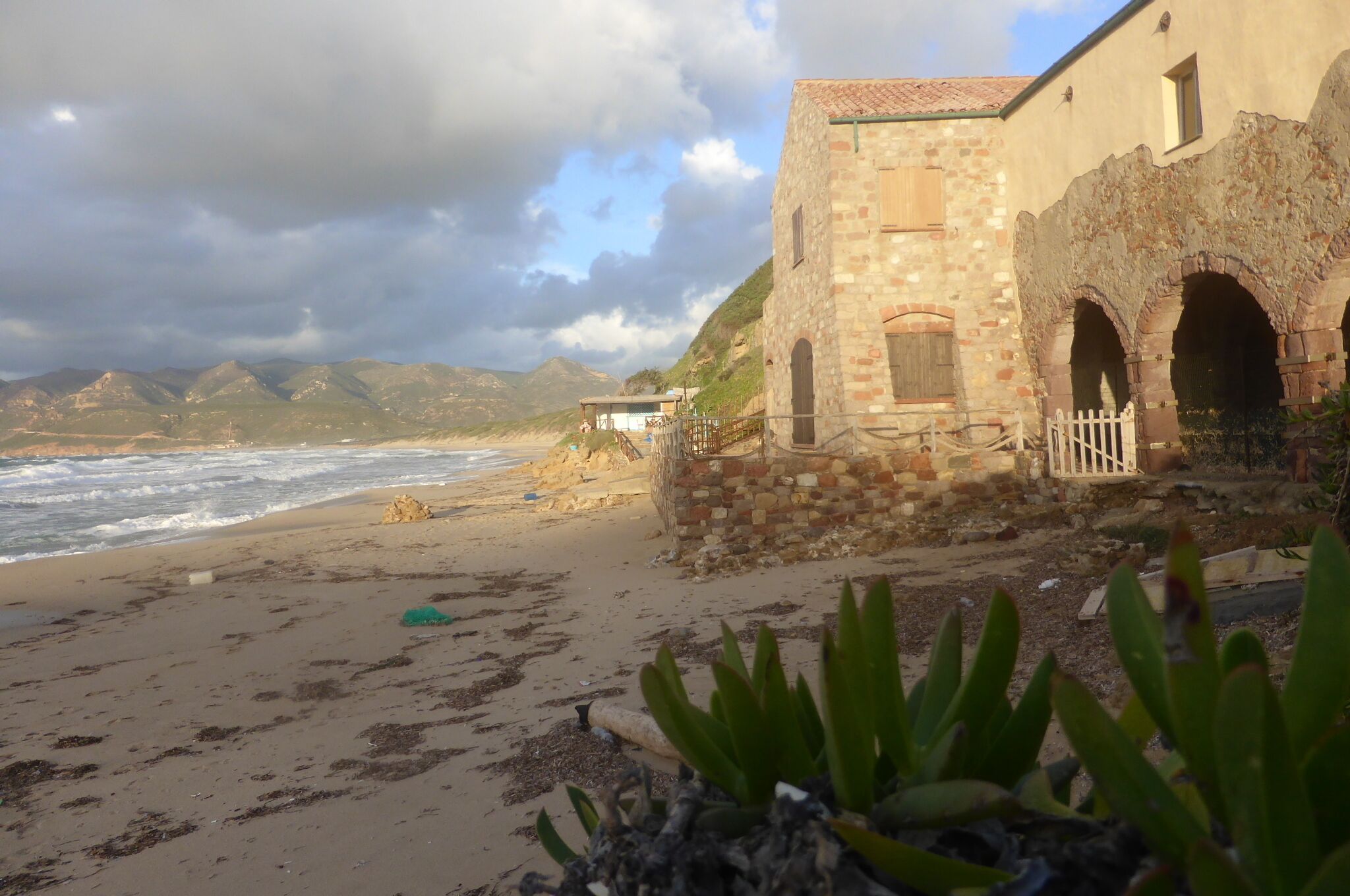 A beachside building with stone walls and arched doorways, overlooking the ocean and mountains.