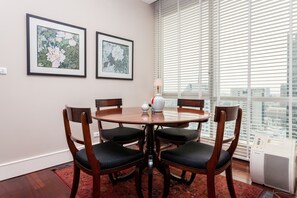 A dining area with a round table, four chairs, two framed floral paintings, a vase, and a window with blinds.