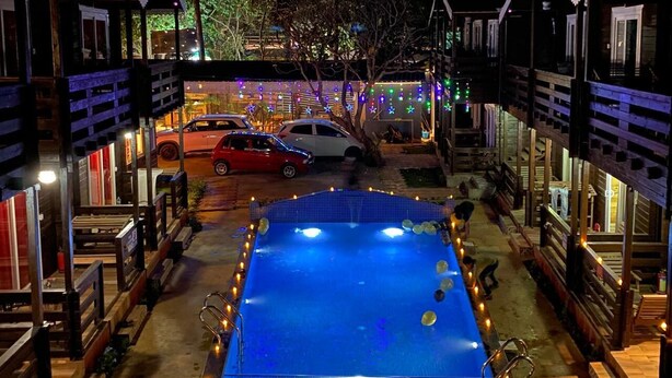 Un espace piscine avec une piscine bleue, entourée d’une terrasse en bois et de bancs, le tout illuminé par des guirlandes lumineuses.