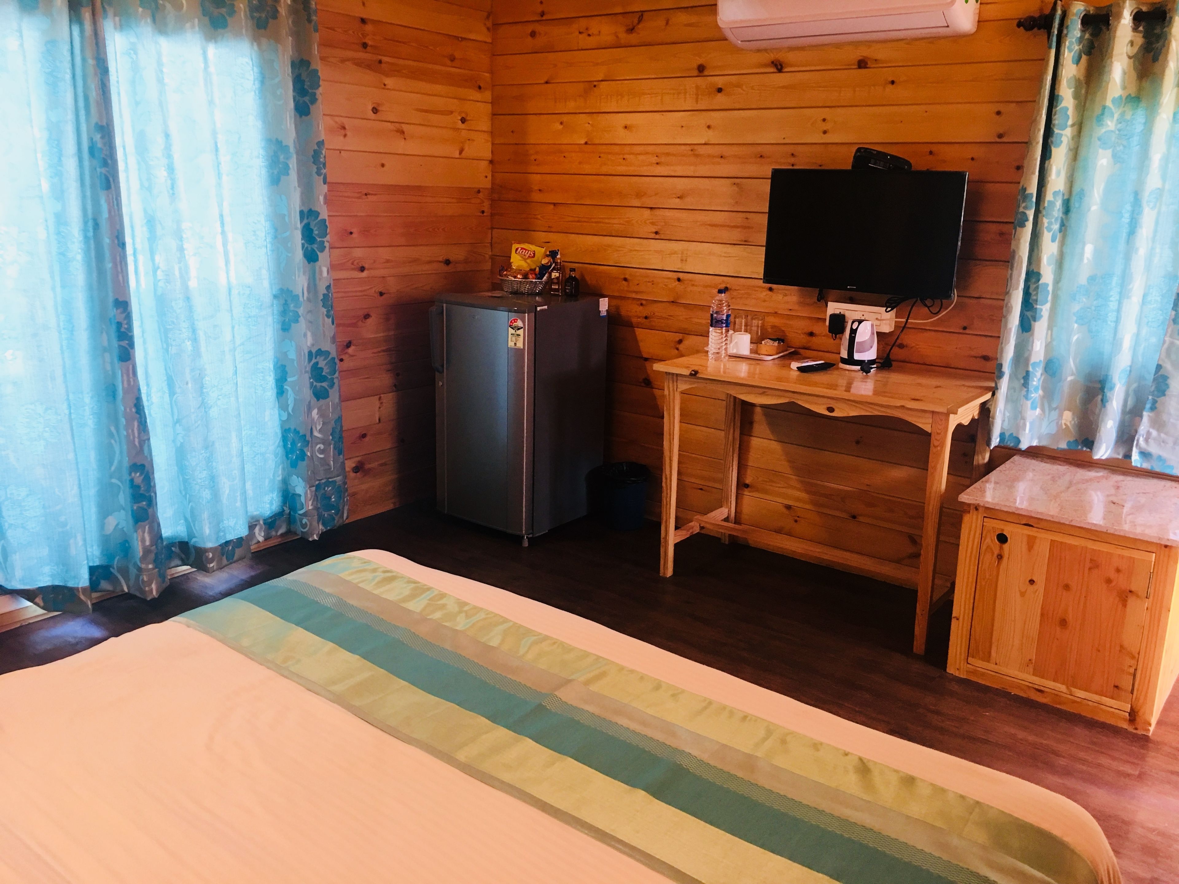 Quarto de cabana de madeira com uma cama, uma escrivaninha, um refrigerador, uma televisão e um pequeno armário.