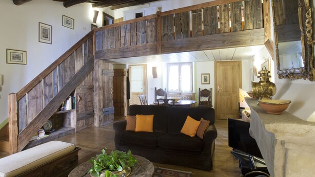 Ein großzügiger Wohnbereich mit einer Holztreppe, einem schwarzen Sofa mit orangefarbenen Kissen und einem rustikalen Kamin.