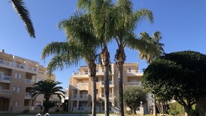 Un área de piscina con palmeras, cielo azul despejado y un edificio de varios pisos con balcones.
