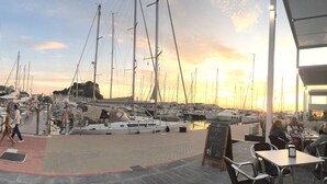 Un puerto deportivo con barcos amarrados, un café con terraza y un atardecer al fondo.