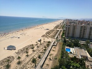 A coastal beach with a long promenade lined with palm trees and a pool area adjacent to a high-rise building.