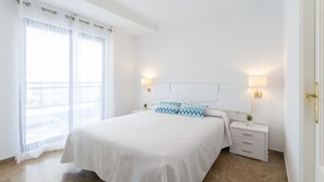 A bedroom with a large bed, two bedside lamps, a nightstand, a window with sheer curtains, and a view of the outside.