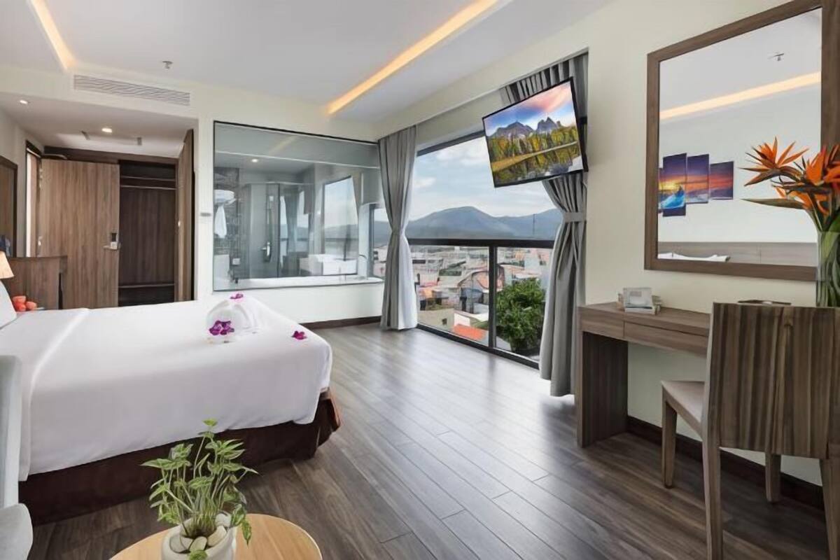 A hotel room with a bed, a desk, a chair, a TV, and a large window with a city view.
