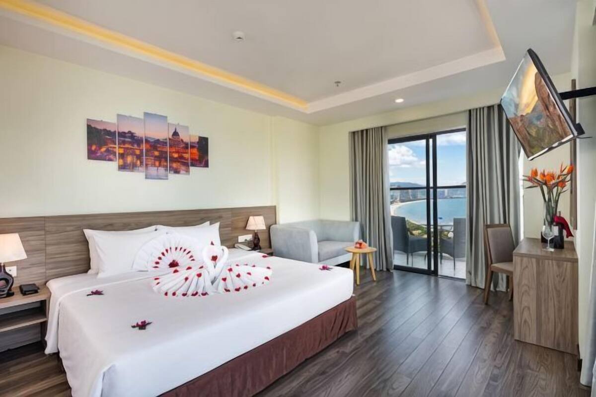 A hotel room with a large bed, a desk, a chair, a TV, and a view of the sea.