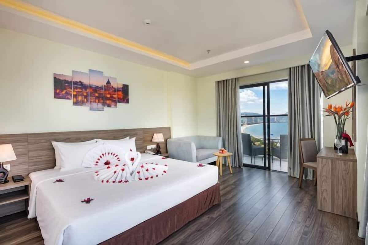 A hotel room with a large bed, a desk, a chair, a TV, and a view of the sea.