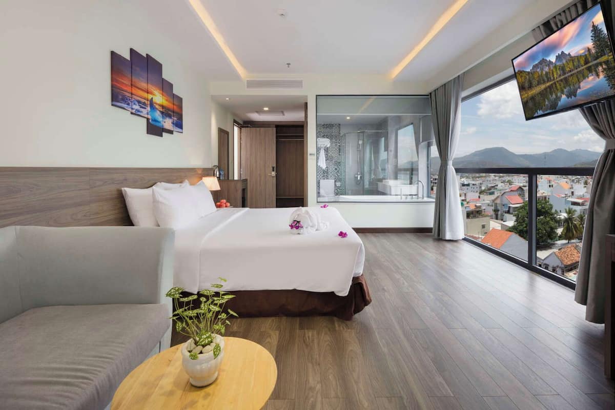 A hotel room with a bed, a sofa, a small table with a plant, a large window with a city view, and a TV on the wall.