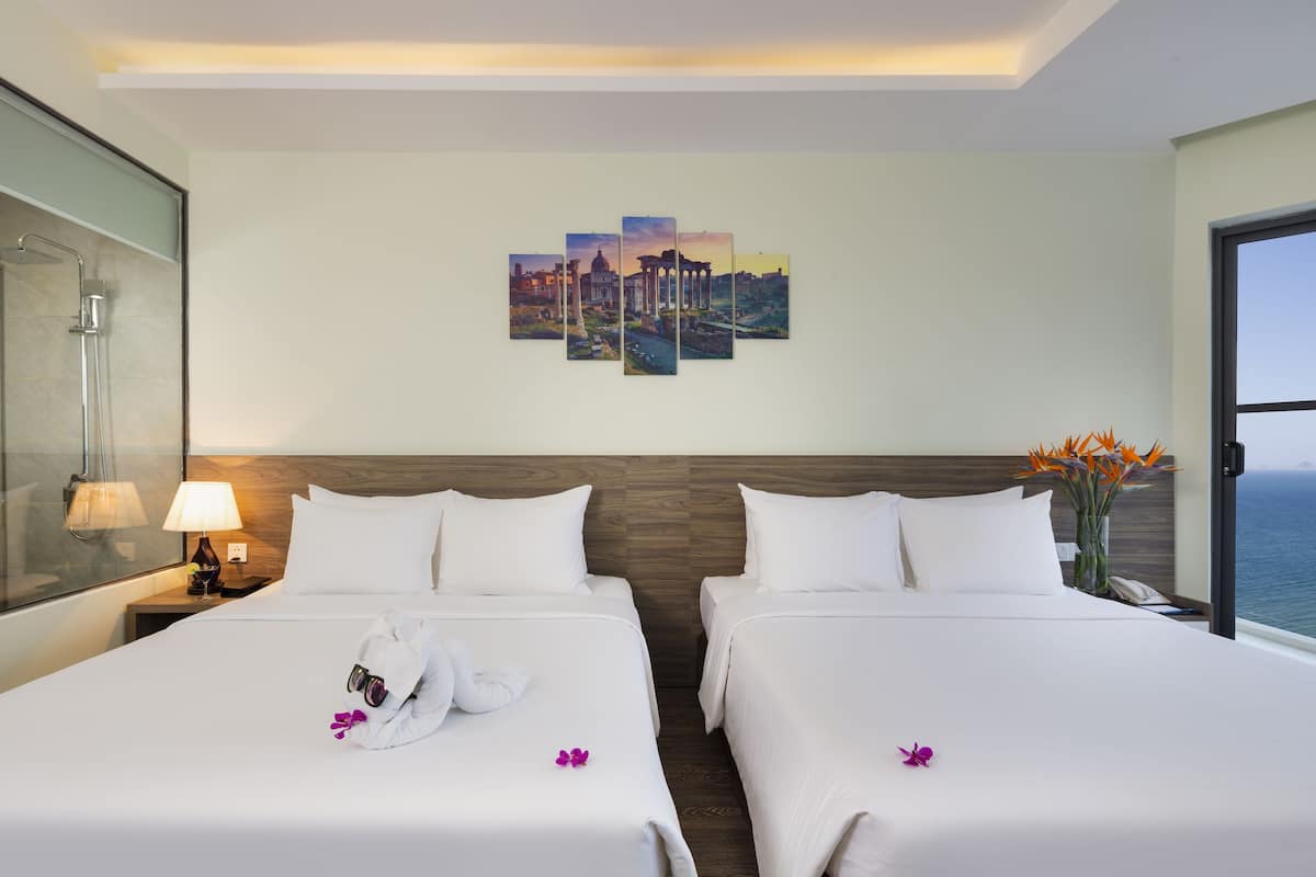 A hotel room with two beds, a glass shower enclosure, a view of the sea, and a cityscape painting on the wall.