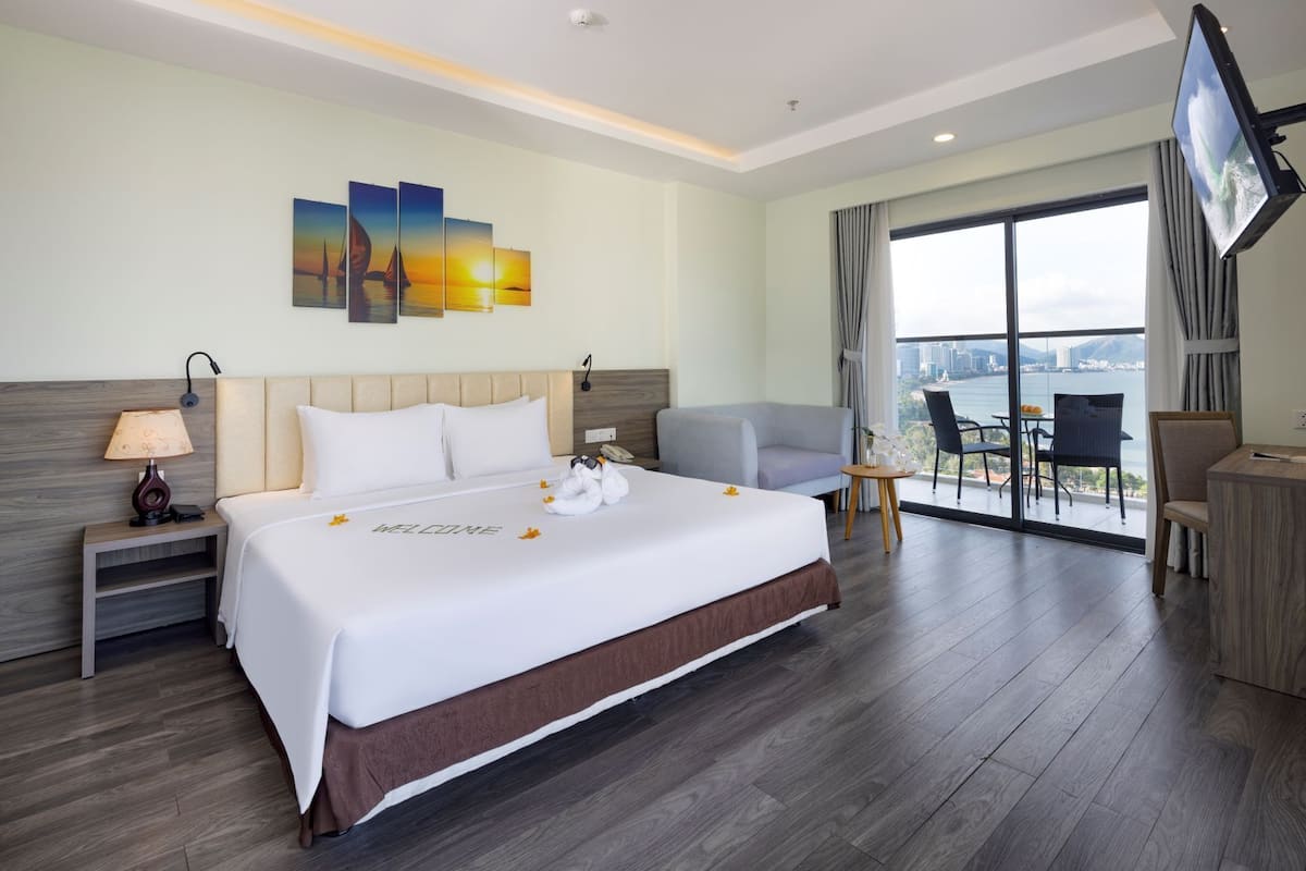 A hotel room with a large bed, a desk with a lamp, a chair, a TV, and a balcony with a view of the city.