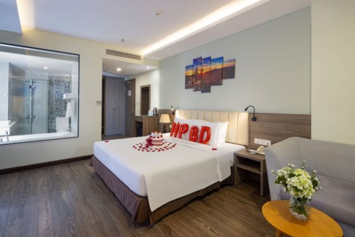 A hotel room with a large bed, a bedside table, a nightstand, a sofa, a small table with flowers, a wall with a sunset painting, and a shower area.