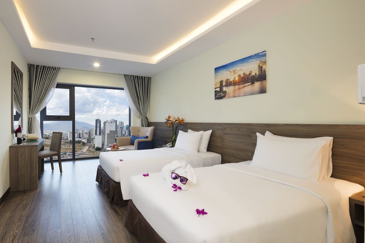 A hotel room with two beds, a desk, a chair, a large window with a city view, and a painting on the wall.
