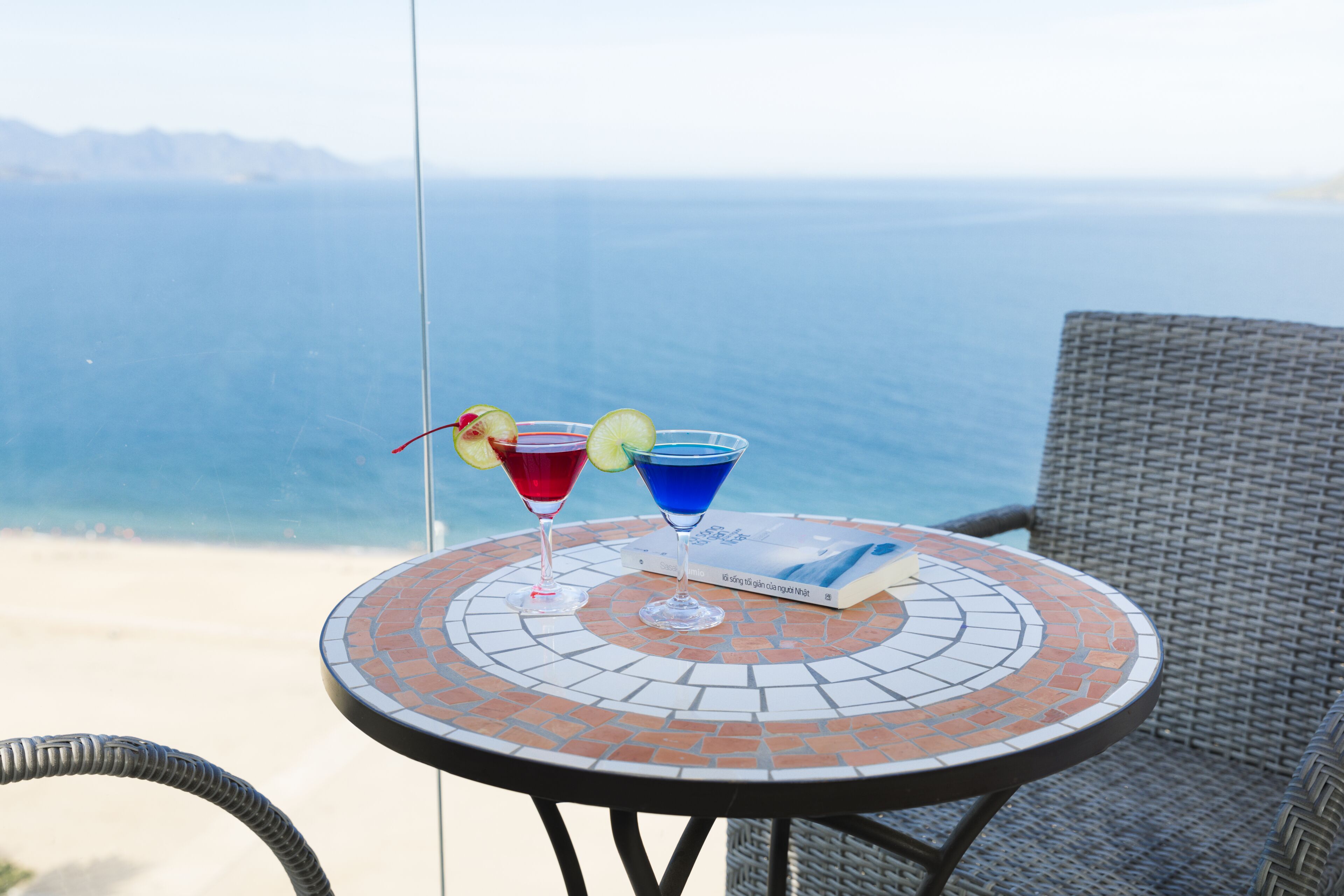 A round table with a mosaic design, two cocktails with garnishes, and a book on it, overlooking a beach and ocean.