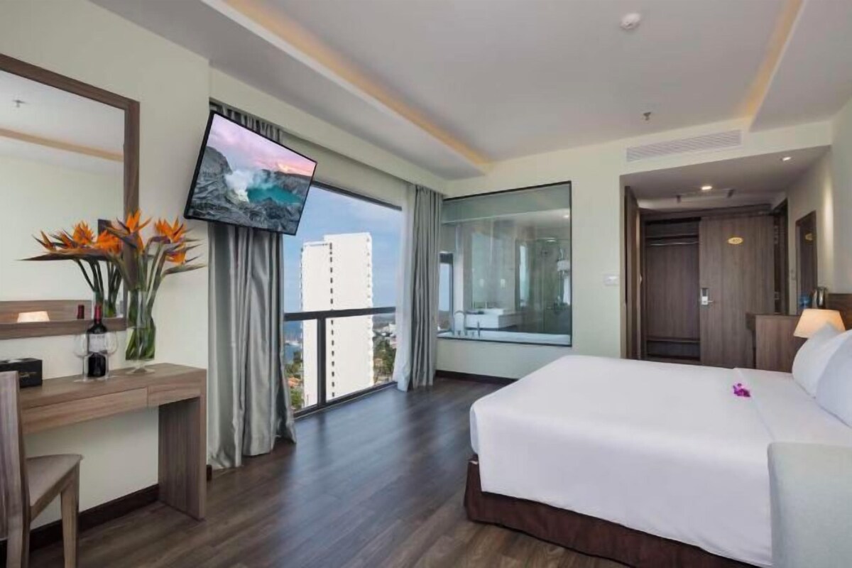 A hotel room with a large bed, a desk with a chair, a TV mounted on the wall, and a window with a view of buildings.