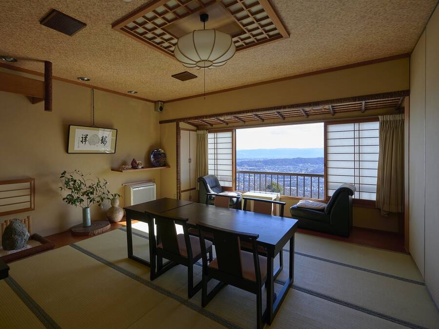 Una habitación tradicional japonesa con suelo de tatami, una mesa baja con sillas, vistas de la ciudad a través de puertas correderas shoji y un ventilador de techo.